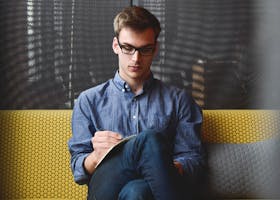 A young man in glasses writes in a notebook while sitting on a stylish couch indoors.